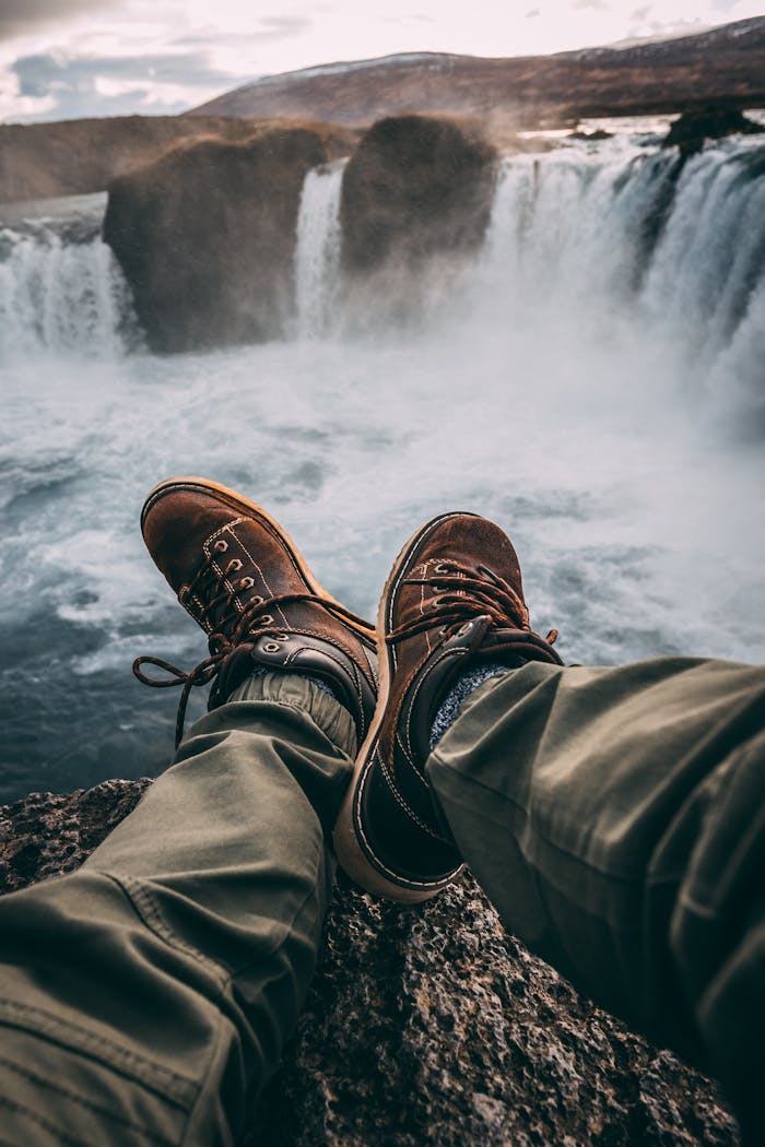 Person in hiking boots sitting at cliff's edge, overlooking breathtaking waterfall.